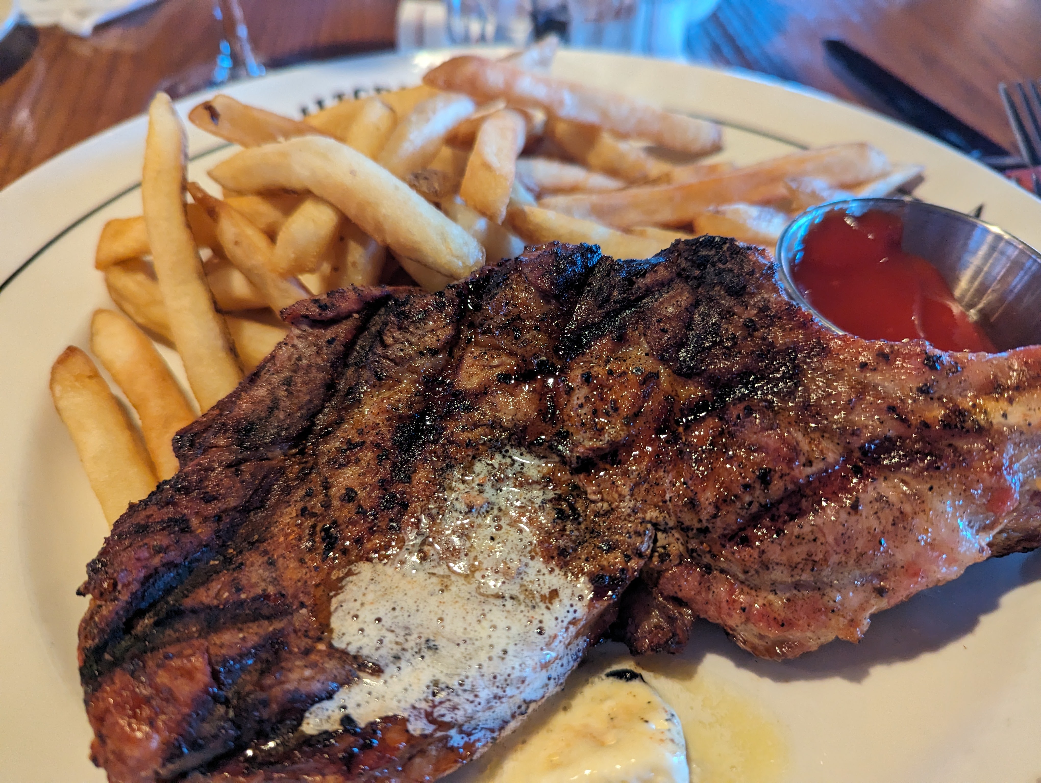 White plate with steak with melted butter and french fries and a small round silver cup of ketchup