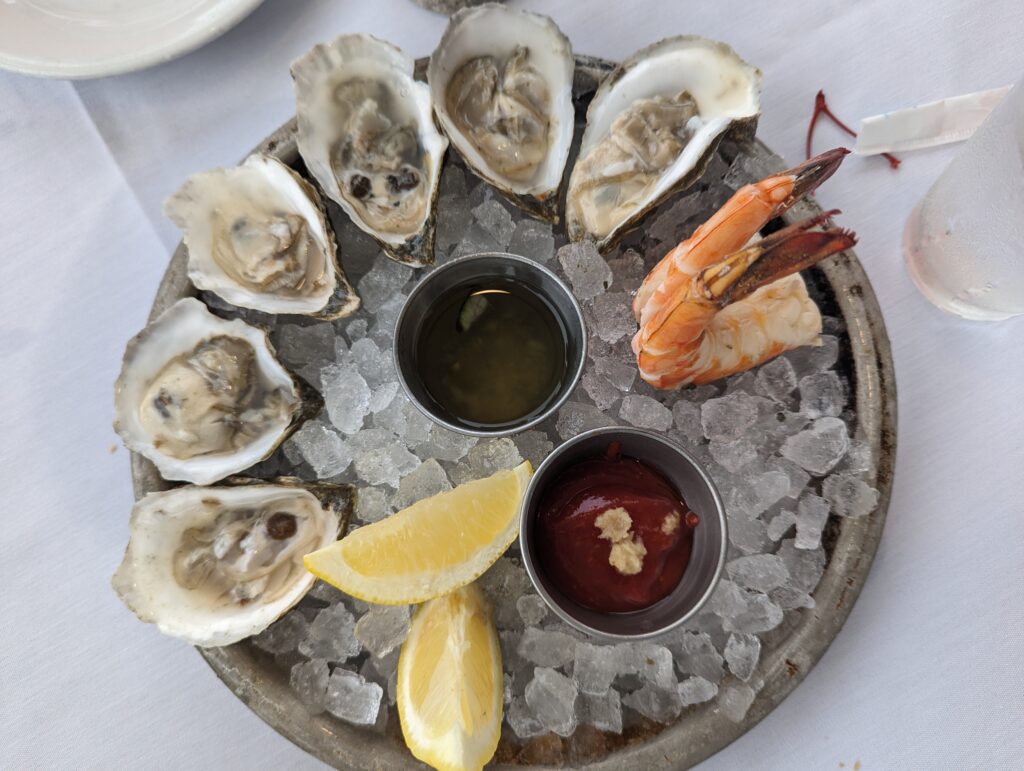round silver platter with ice and two jumbo pink shrimp and 6 oysters on the half shell with red cocktail sauce, a vinegrette, and 2 lemon wedges