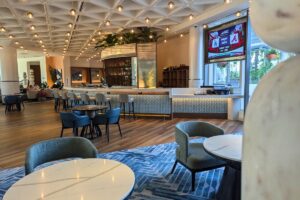 hardwood floors with blue, chevron print rugs, with round white marble tables and blue chairs. There are round lights that dot the ceiling and a curved bar with light blue barstools